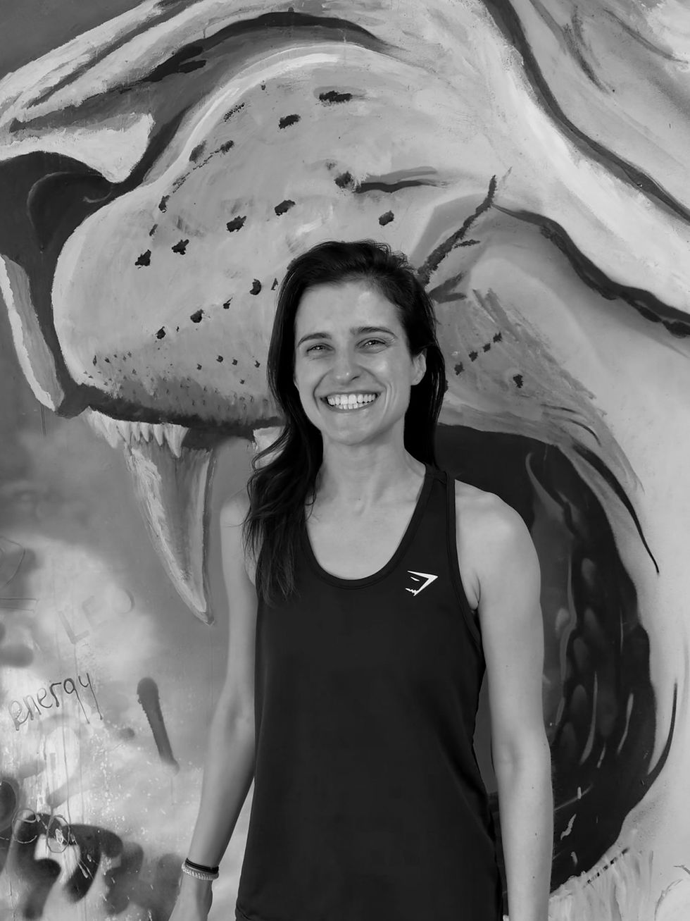 Smiling woman in a black tank top stands before a black-and-white whale mural with faint graffiti reading energy.