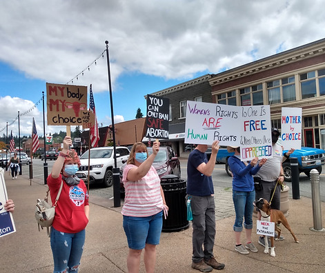 A group stands on a corner in downtown Monroe demonstrating for reproductive rights. Signs read: "My body my choice," "You can only ban safe abortion," "women's rights are human rights," "no one is free when others are oppressed," and "mother by choice." A dog, a boxer, has a sign around their neck saying "Heck the Patriarchy"