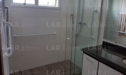 Imagem de um banheiro claro onde se vê um amplo box, com barras de apoio brancas