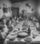 Dieses Bild zeigt eine harmonische Familienszene, bei der mehrere Generationen um einen großen Esstisch versammelt sind. Das Essen steht im Mittelpunkt des Zusammenseins, und die Atmosphäre erinnert an traditionell ländliche italienische Familien. Die gedeckte Tafel ist mit einfachen, aber reichhaltigen Speisen wie Brot, Pasta und Salaten bestückt. Die dunklen, rustikalen Wände und das schlichte Mobiliar lassen vermuten, dass sich die Szene in einem bescheidenen ländlichen Zuhause abspielt. Eine Frau, vermutlich die Großmutter oder Matriarchin, steht aufrecht, während sie das Essen serviert. Die Anwesenden, darunter Kinder und Erwachsene, wirken entspannt und genießen das gemeinsame Mahl. Es vermittelt ein starkes Gefühl von Gemeinschaft, Tradition und familiärer Wärme.