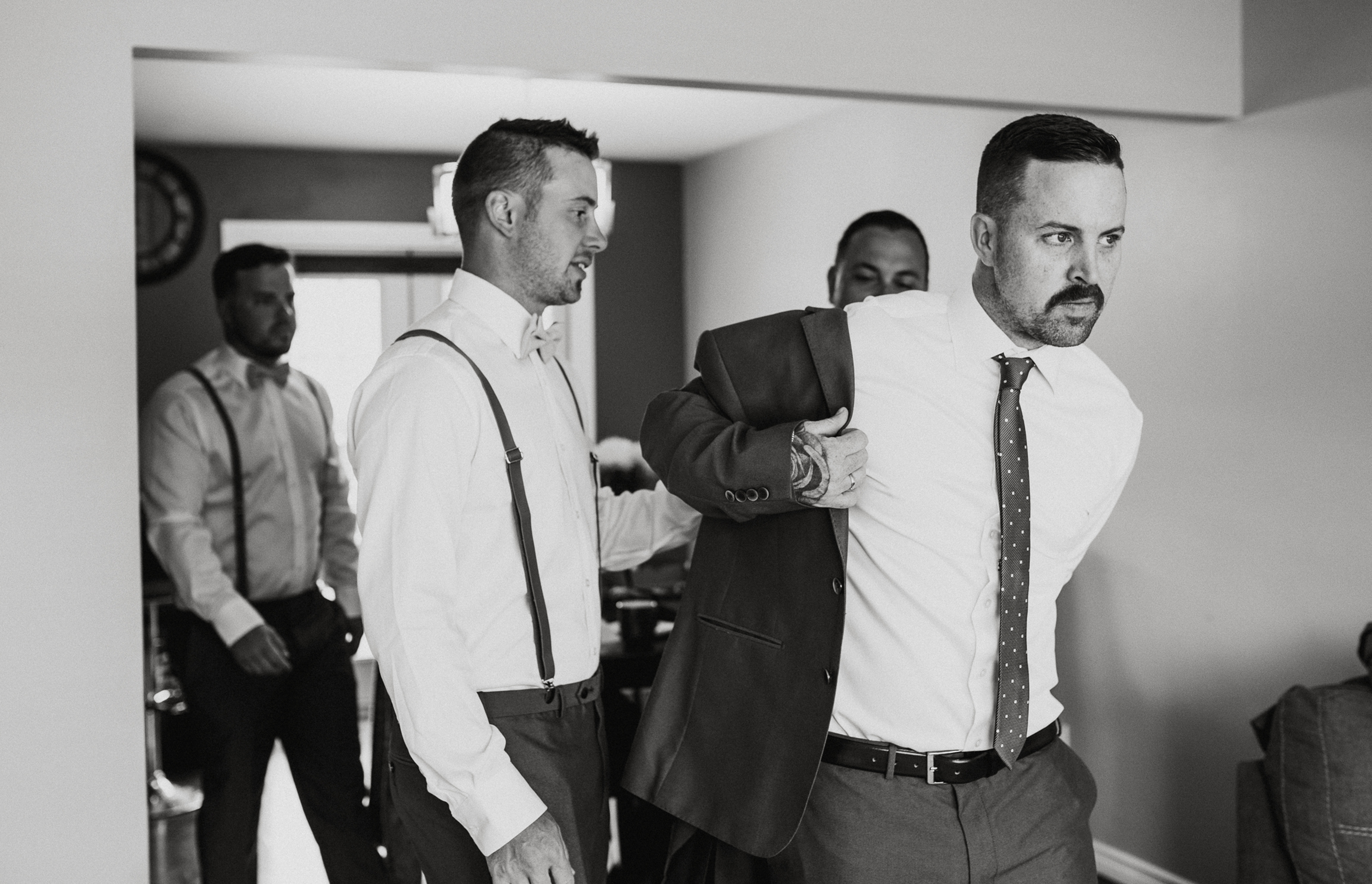 Second photographer captures groomsmen in dress shirts and suspenders help one man put on a suit jacket indoors, black-and-white, focused pre-event mood