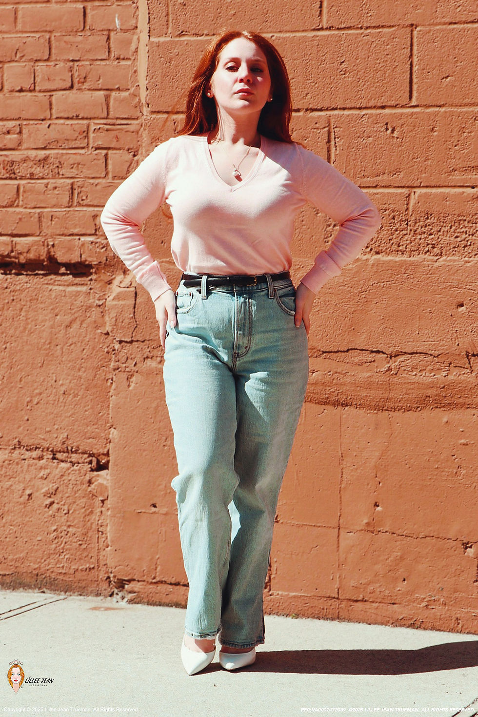 Lillee Jean Trueman, the actress, in an official portrait, wearing a pink jumper, blue denim, and white shoes. Natural redhair is flowing down with earrings.