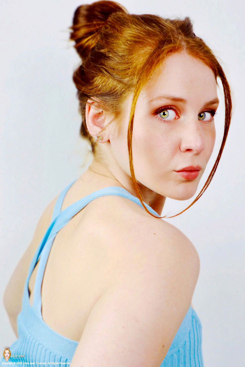 Official celebrity portrait of actress Lillee Jean Trueman. Her striking blue-green eyes are the focus, complemented by a blue shirt and her signature red hair, styled in an updo.