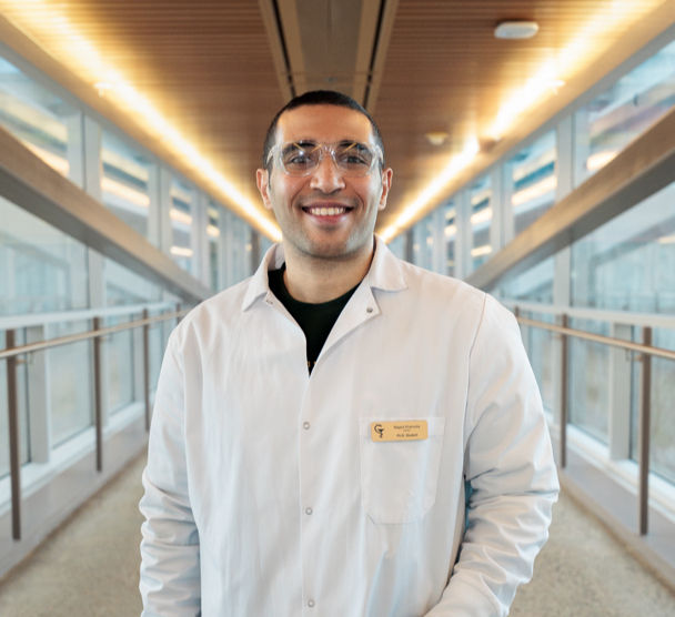 A young man poses in a well lit skywalk, wearing a lab coat and safety goggles.