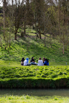 A group of young people are sitting in a circle on a grassy lawn. Trees are in the background, it's early Spring so they have no leaves on.