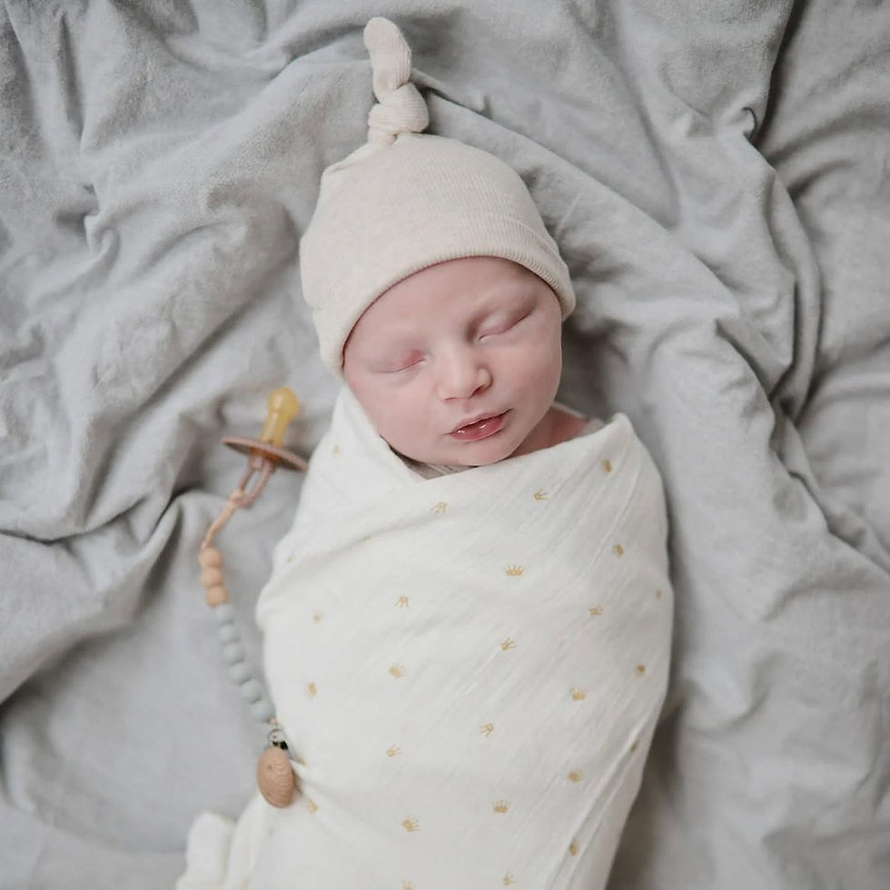 Sleeping newborn swaddled in a white Mushie muslin cloth with a gold crown print, wearing a beige knotted beanie on a grey be