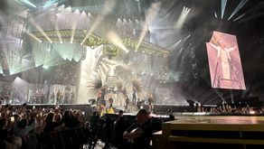 Robbie Williams performing on stage in Kaunas at Darius and GirÄ—nas Stadium, with stage lights and fans in the foreground