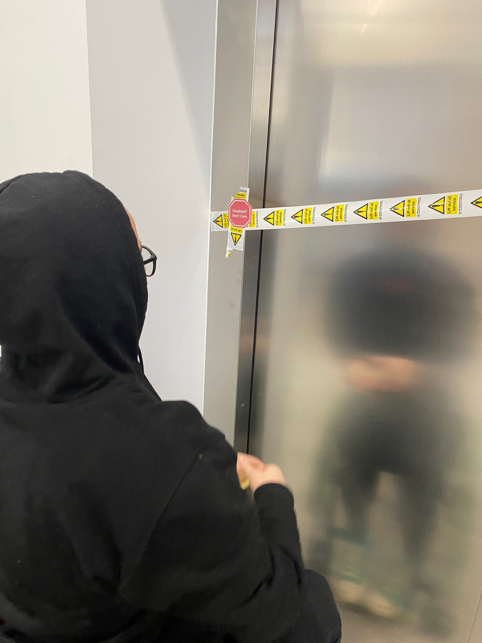 A person in a black hoodie sticks a red octagon sticker that says “Disabled? Don’t care” to “lift out of order” tape on a lift’s doors