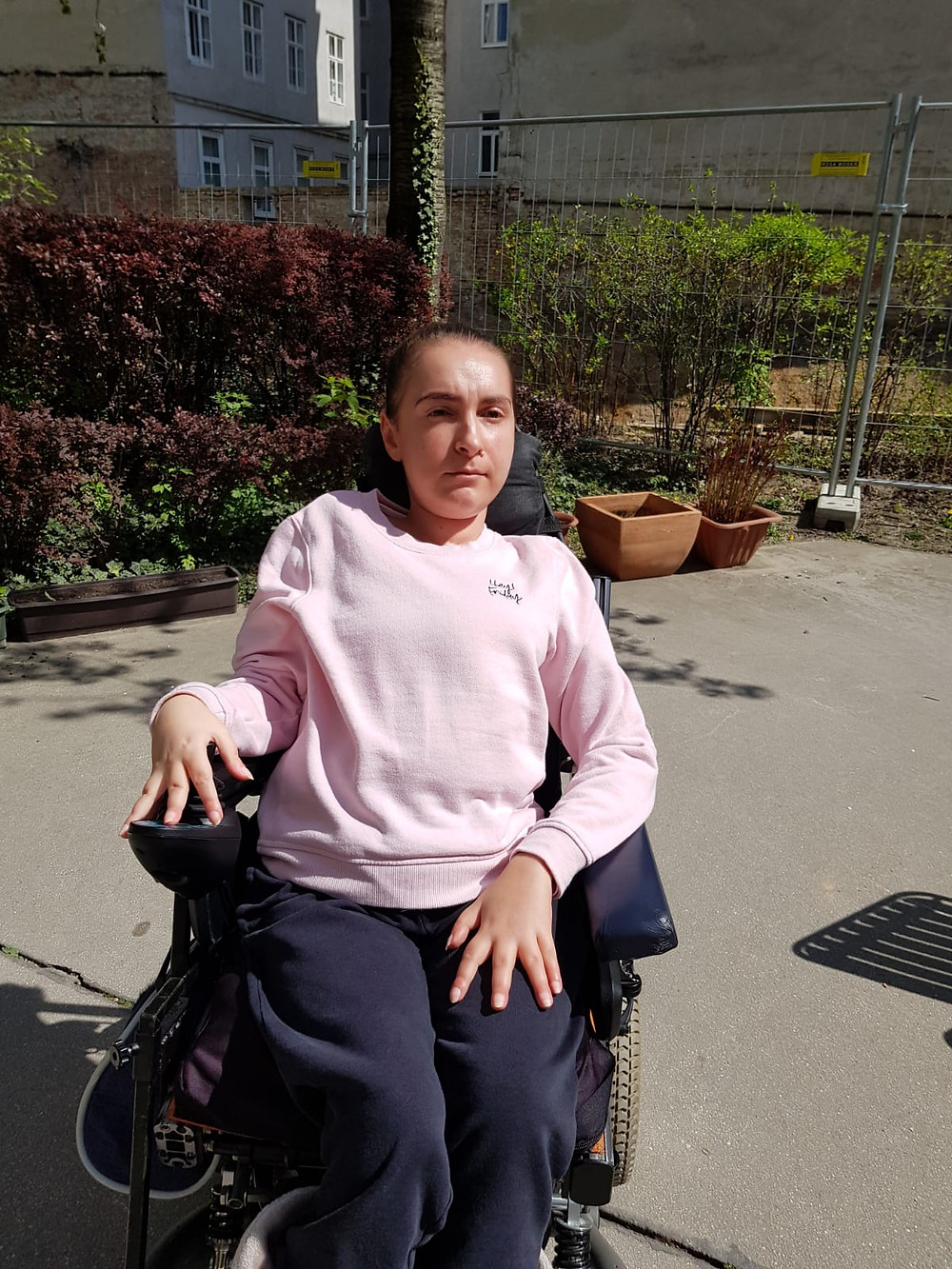 Lidija Ljubic in einem sonnigen Vorhof im Rollstuhl sitzend mit rosa Pullover
