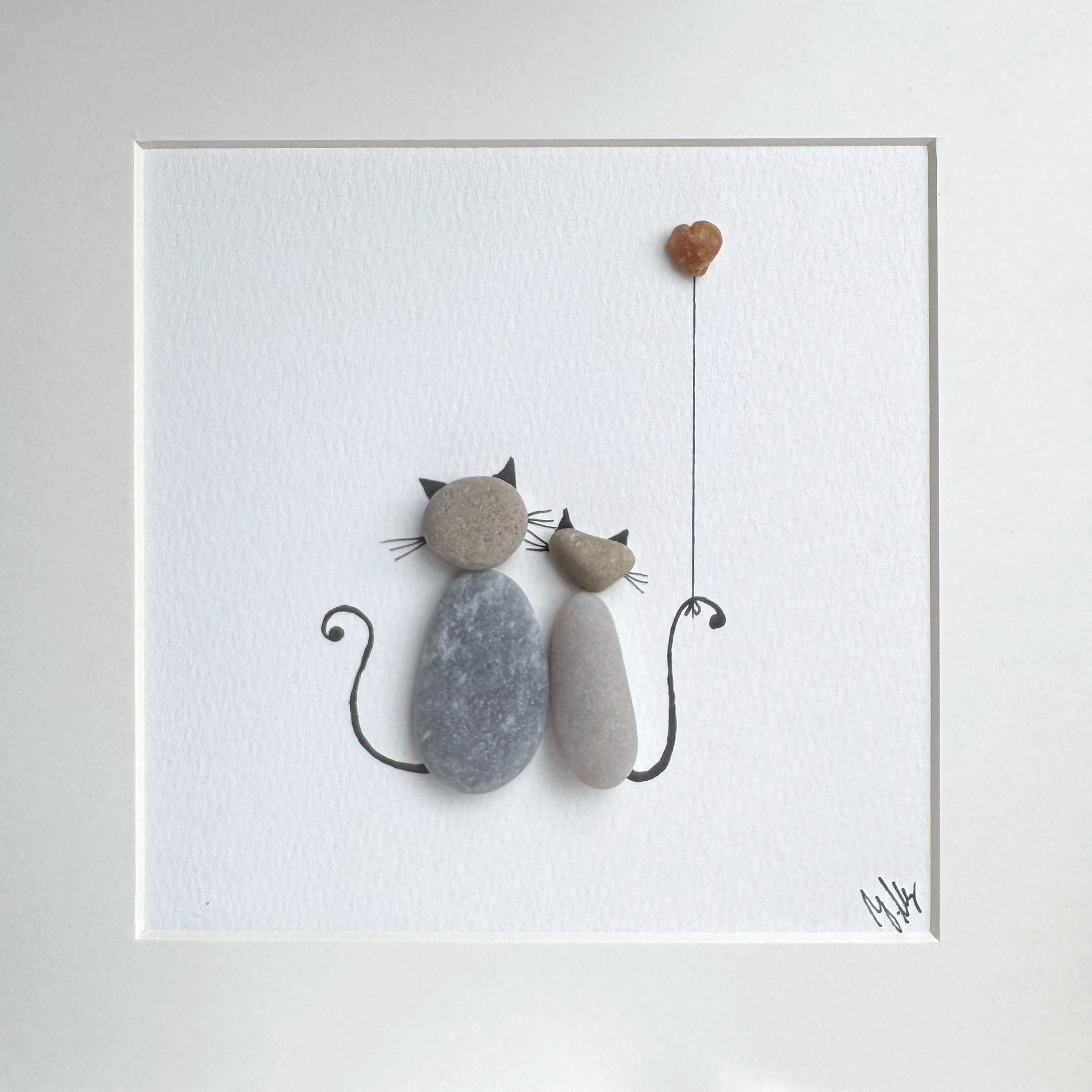 Handgefertigtes Kieselsteinbild mit zwei Katzen, die eng aneinandergekuschelt unter einem herzförmigen Steinballon sitzen.
