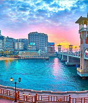 Vue panoramique du pont Stanley à Alexandrie au coucher du soleil, avec des tours emblématiques et des reflets colorés sur l’eau. Scène urbaine animée avec une architecture remarquable.