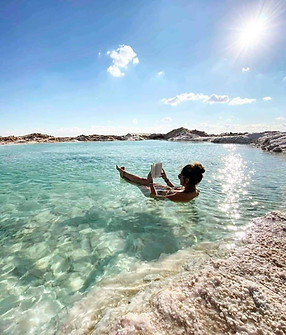 Une personne flottant dans une eau turquoise claire, lisant un livre, avec un paysage rocheux en arrière-plan et un soleil éclatant au-dessus.