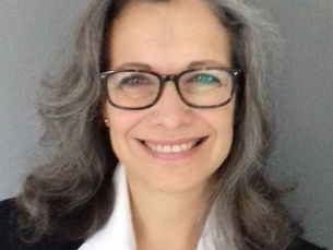 Woman with glasses smiling, wearing a black blazer and white shirt, against a gray background. Gray hair and cheerful expression.