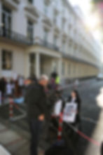 People holding "LONG COVID KIDS" signs protest outside a white building. A camera crew films, with bright sunlight in the background.
