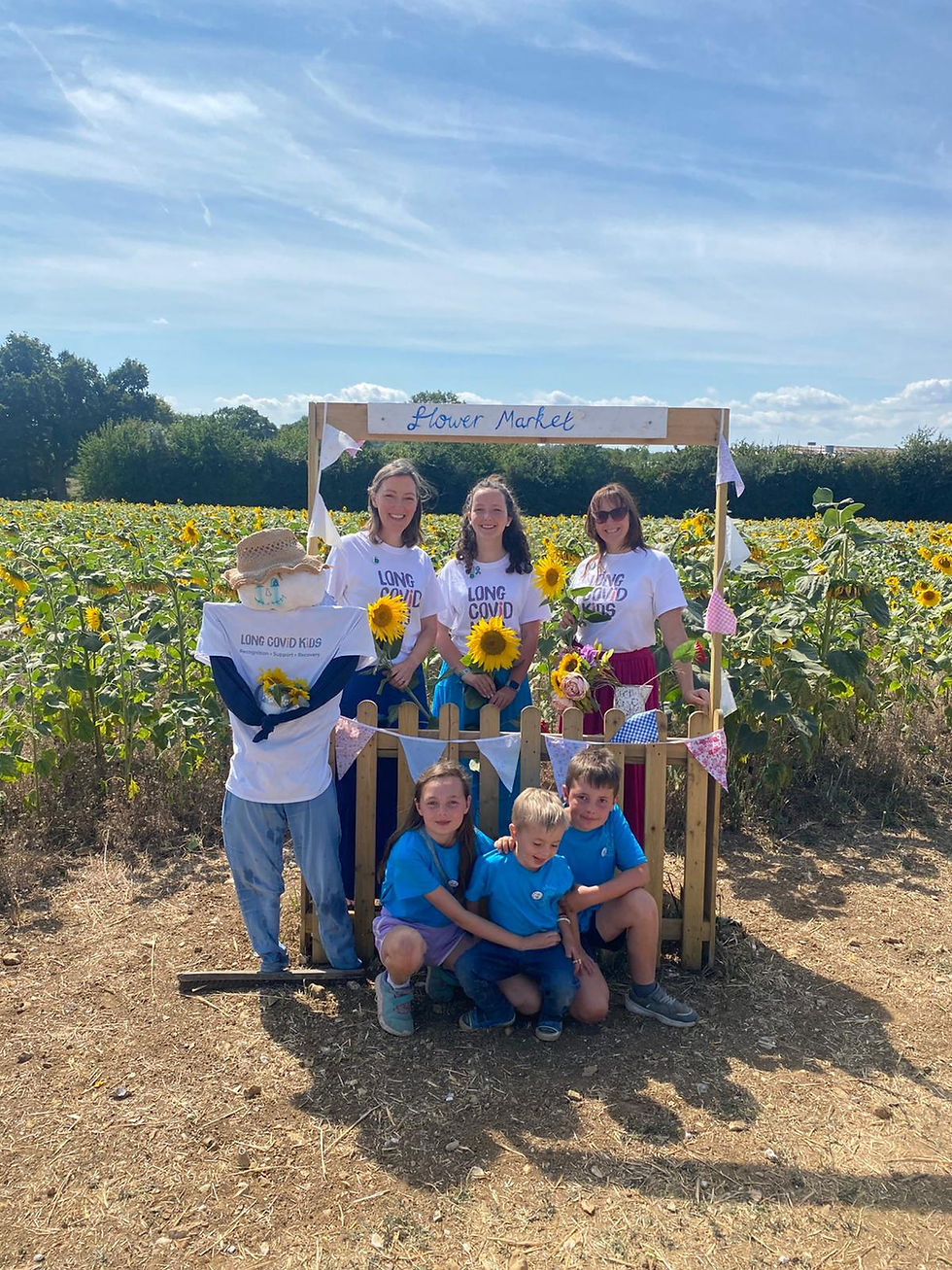 People in "Long Covid Kids" shirts hold sunflowers at a flower market stand in a sunny sunflower field. A scarecrow and children are present.