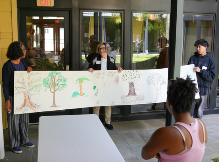 Three people hold a large drawing of trees indoors, observed by a seated person. The setting is bright with large windows.