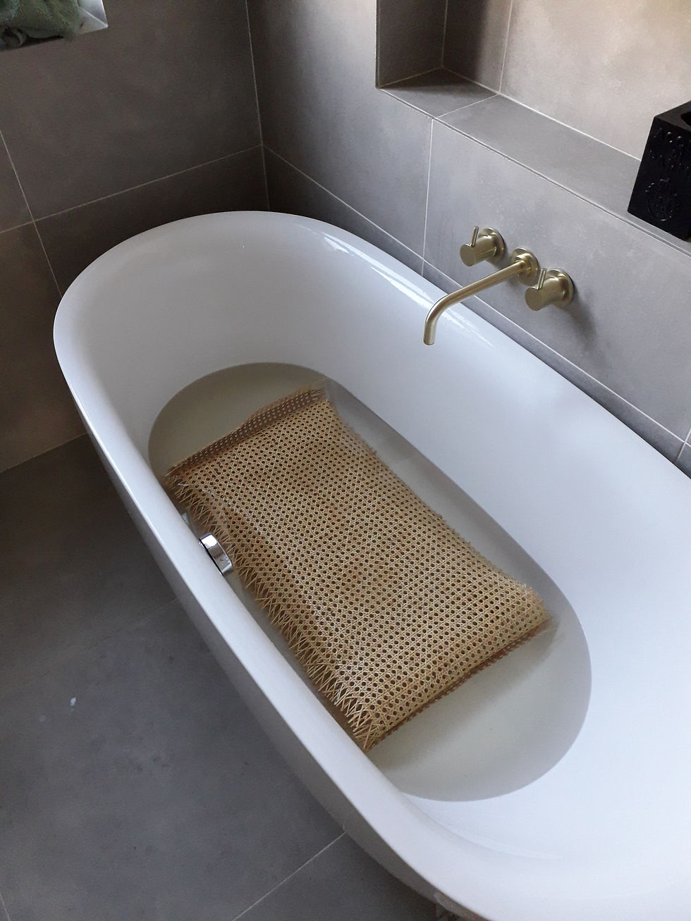 Rattan weave soaking in white bathtub with water. Brass faucet on gray tiled wall. Minimalist and serene bathroom setting.