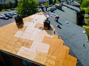 Roofers replace shingles on a large apartment roof, with plywood exposed, tools and materials scattered, and parked cars below.