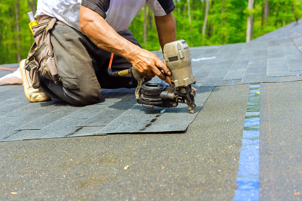 Worker using a nail gun to install roof shingles on a rooftop, with green trees blurred in the background.