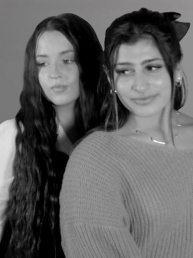 a black and white photo of two young women standing close together, looking off camera. Each has distinct hairstyles: one with long, wavy hair, and the second with a ponytail. They are posing together for a professional photo shoot.