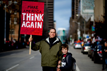 A father and son stand in a street holding a red sign reading "Make America Think Again"