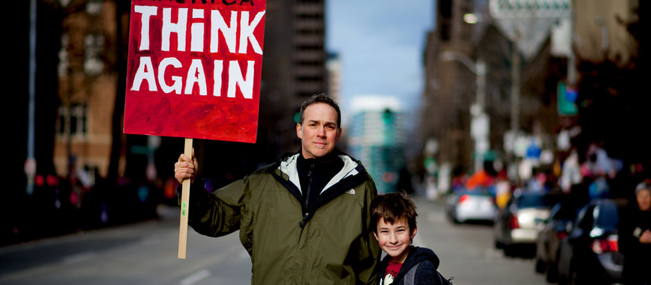 A father and son stand in a street holding a red sign reading "Make America Think Again"