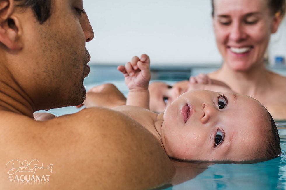 Baby floating in a pool on a man's shoulder, with a smiling woman in the background. Water reflects soft light, creating a calm scene.
