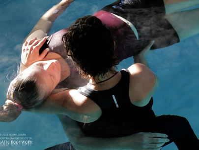 Two people in a pool, one supporting the other who is floating serenely. Water reflects light, creating a tranquil mood. Text reads: Aquatic Bodywork.