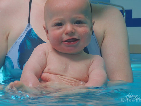 Baby smiling in a pool, held by an adult. Blue swimsuit with leaf pattern. Water droplets visible on baby. Text_ Adaya © AQUANAT_
