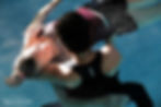 A woman with short dark curly hair with her back to the camera is standing in chest level pool water and gently holding, stretching and embracing another woman who is in a supine floating position and is closing her eyes