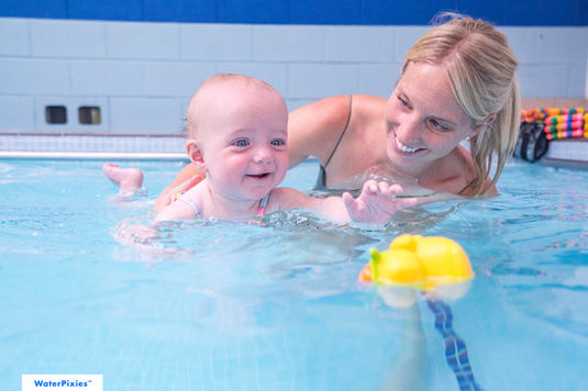 Smiling mother is holding her little baby girl who is smiling and reaching out to grab a toy duck in the pool