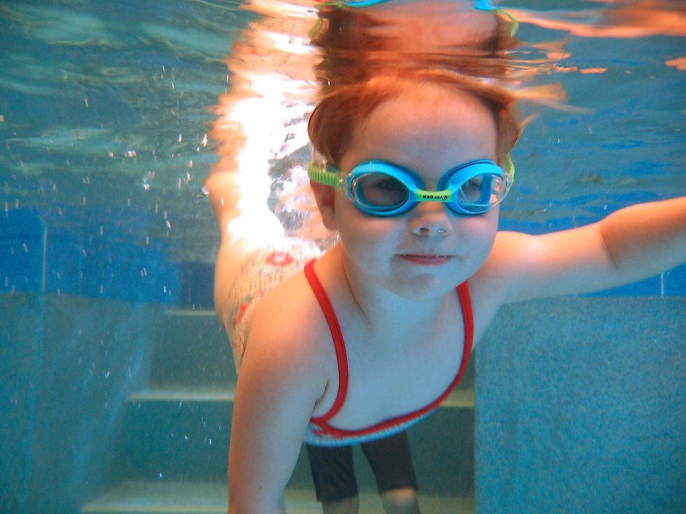 Child wearing blue swim goggles floats underwater in a pool, smiling near the steps and wall, with rippling blue light above.