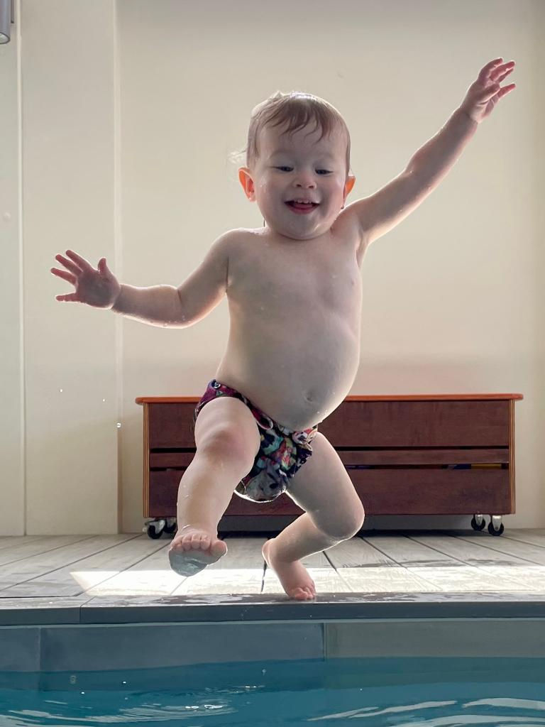Toddler joyfully jumps into a pool, arms outstretched. Wears colorful swim diaper. Indoor setting with wooden bench in the background.