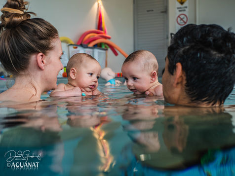 Two babies in a pool with two adults, smiling. Colourful pool noodles in the background create a playful atmosphere.