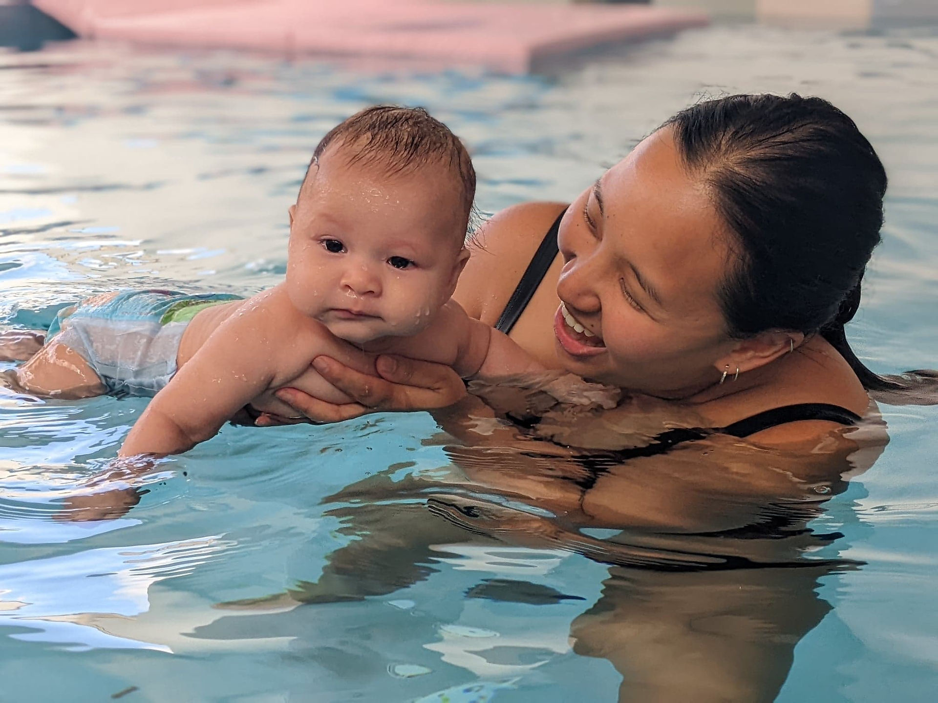 Woman joyfully holds baby swimming in a pool. Clear water surroundings, baby in swim diaper. Text "© Aquanat" visible.