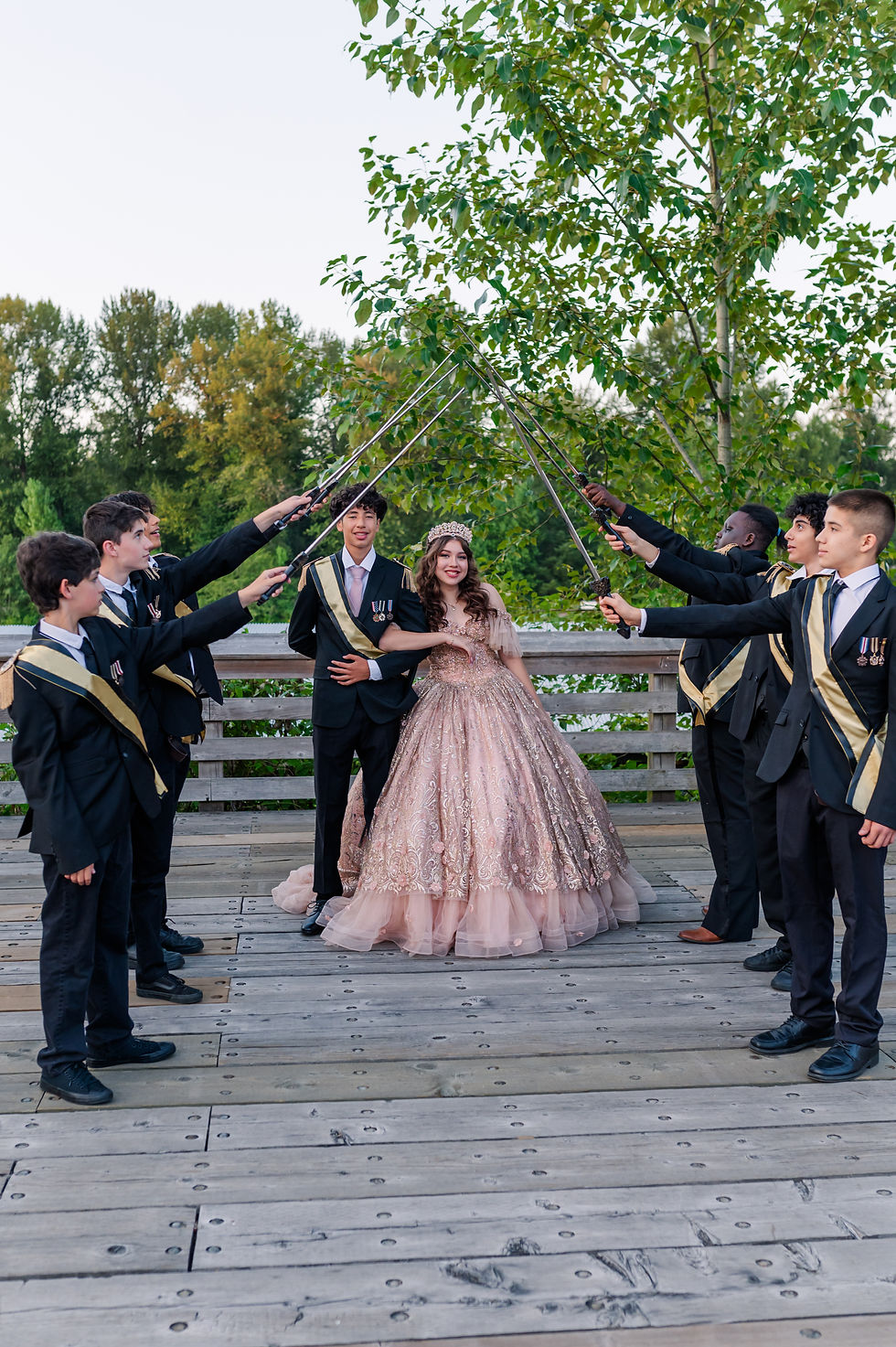 A girl in a pink embellished gown and crown stands under a sword arch held by eight boys in suits on a wooden deck with trees behind.