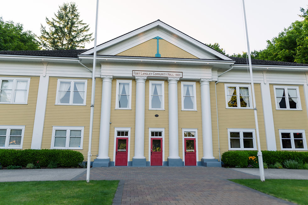 Yellow community hall with white columns and red doors, set in lush greenery. Sign reads "Fort Langley Community Hall 1931."