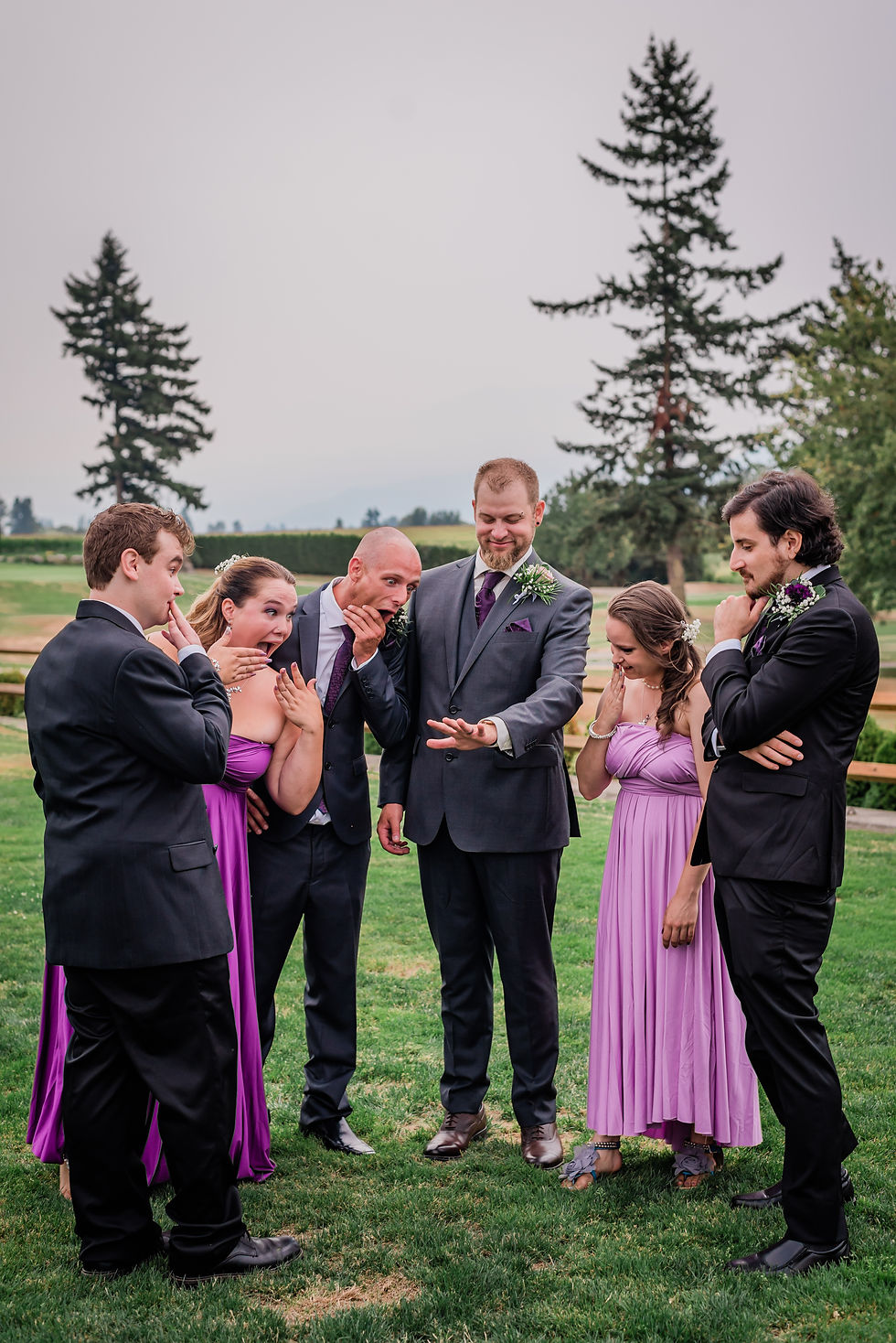 A group in formal wear reacts with surprise to a man's hand, likely showing a ring, in a park with trees and grass.