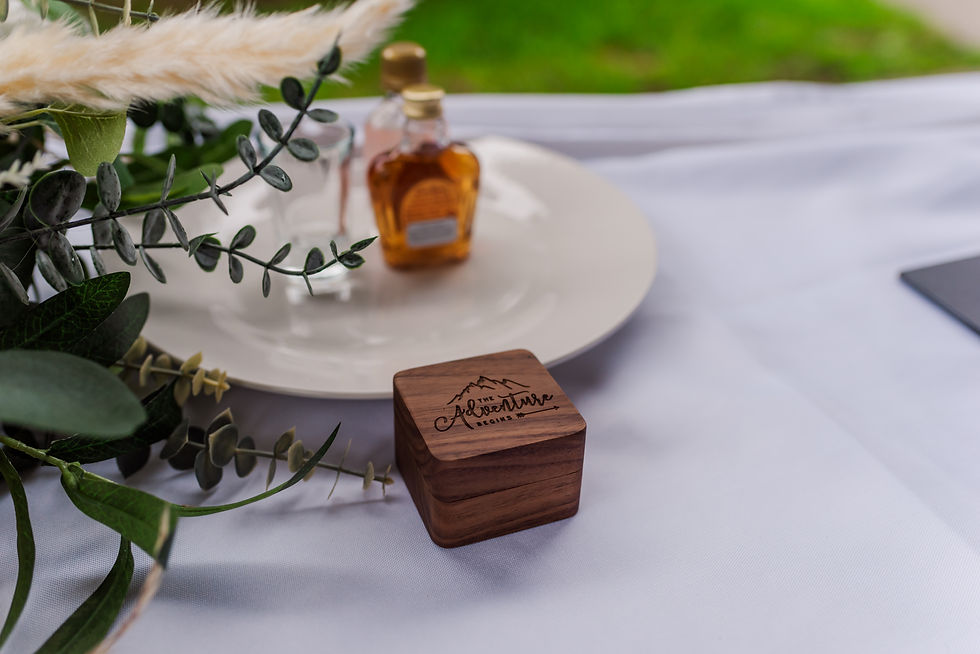 Wooden box with "The Adventure Begins" text, near a white plate with a small bottle and greenery, on a white tablecloth. Outdoor setting.