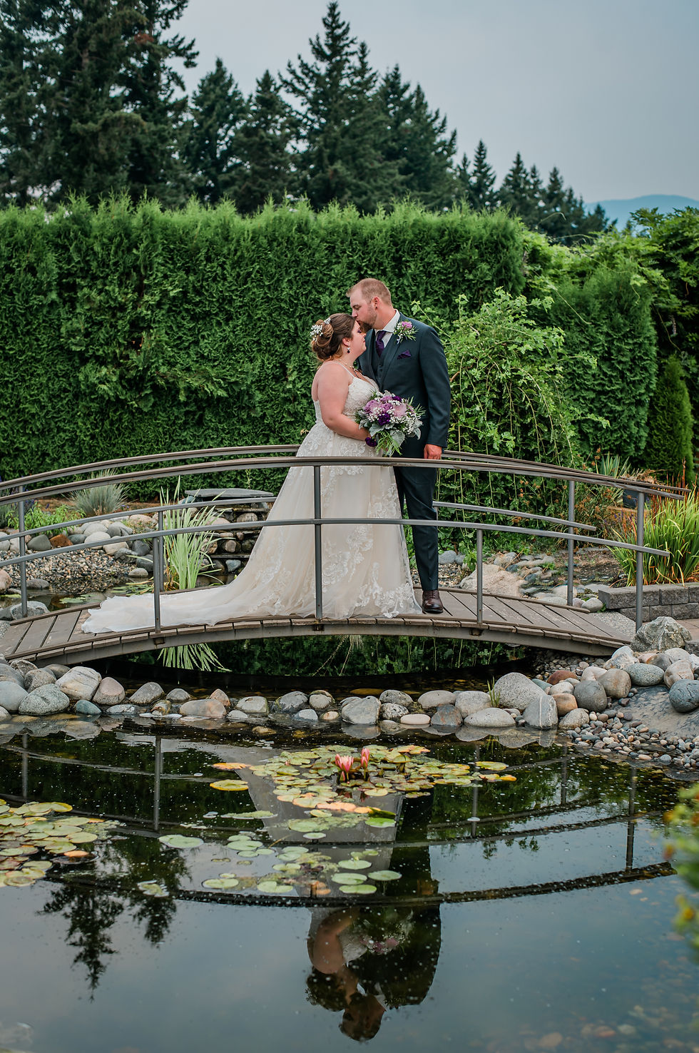 Bride in white gown and groom in dark suit kiss on a small bridge. Lush greenery, pond, and reflection create a romantic setting.