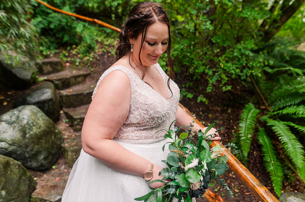 Bride in a beaded gown holds a bouquet, standing on stone steps in a lush green garden, looking down and smiling softly.