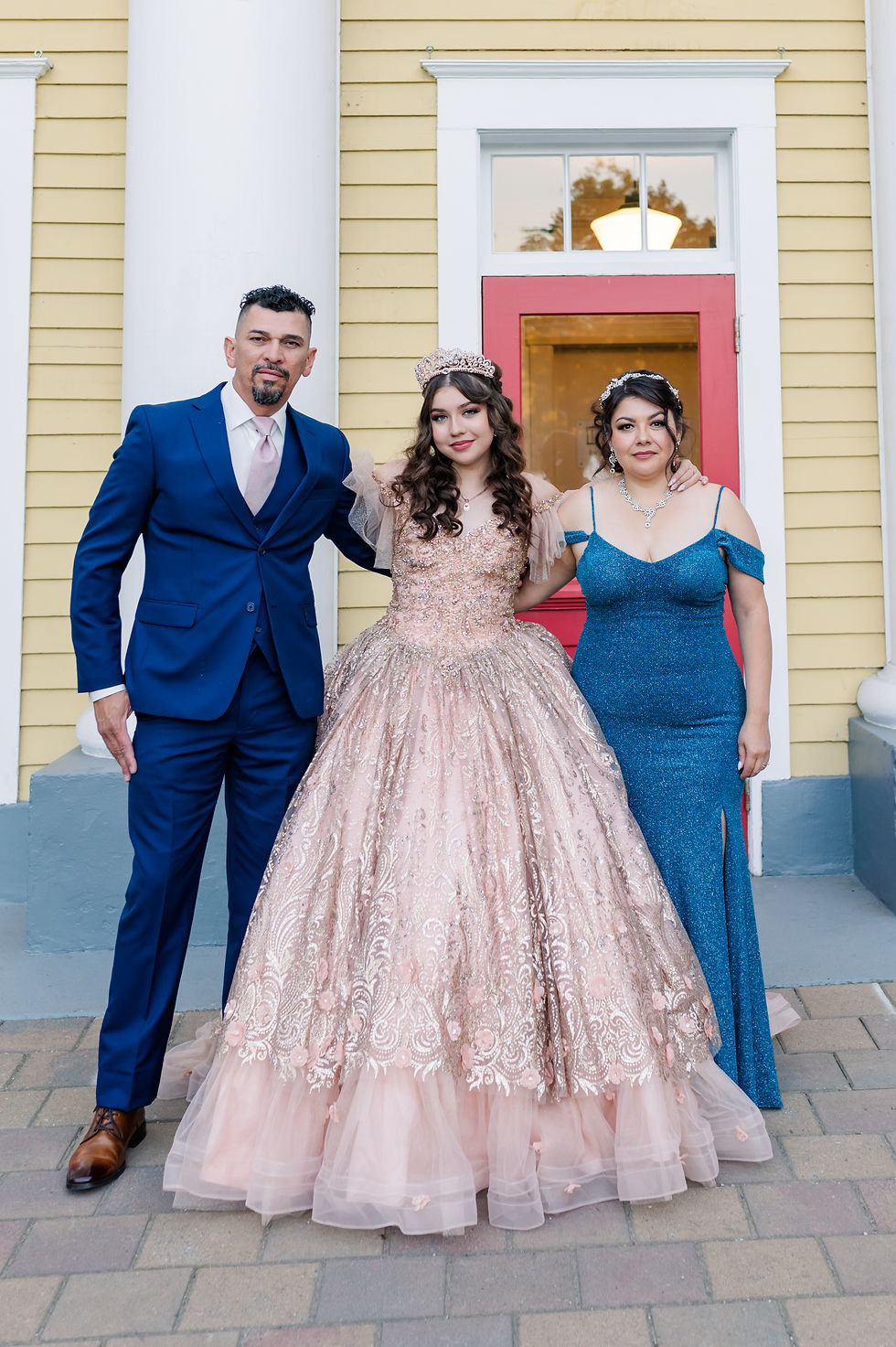 Three people pose in formal attire; a man in a blue suit, a woman in a pink gown with a crown, and another in a blue dress. Yellow house backdrop.
