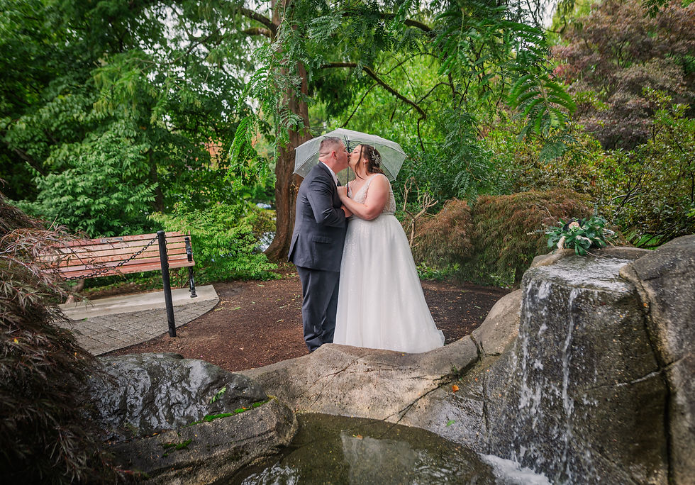 A couple kisses under an umbrella in a lush garden. Bride in white gown, groom in suit. Nearby waterfall and bench add to romantic scene.