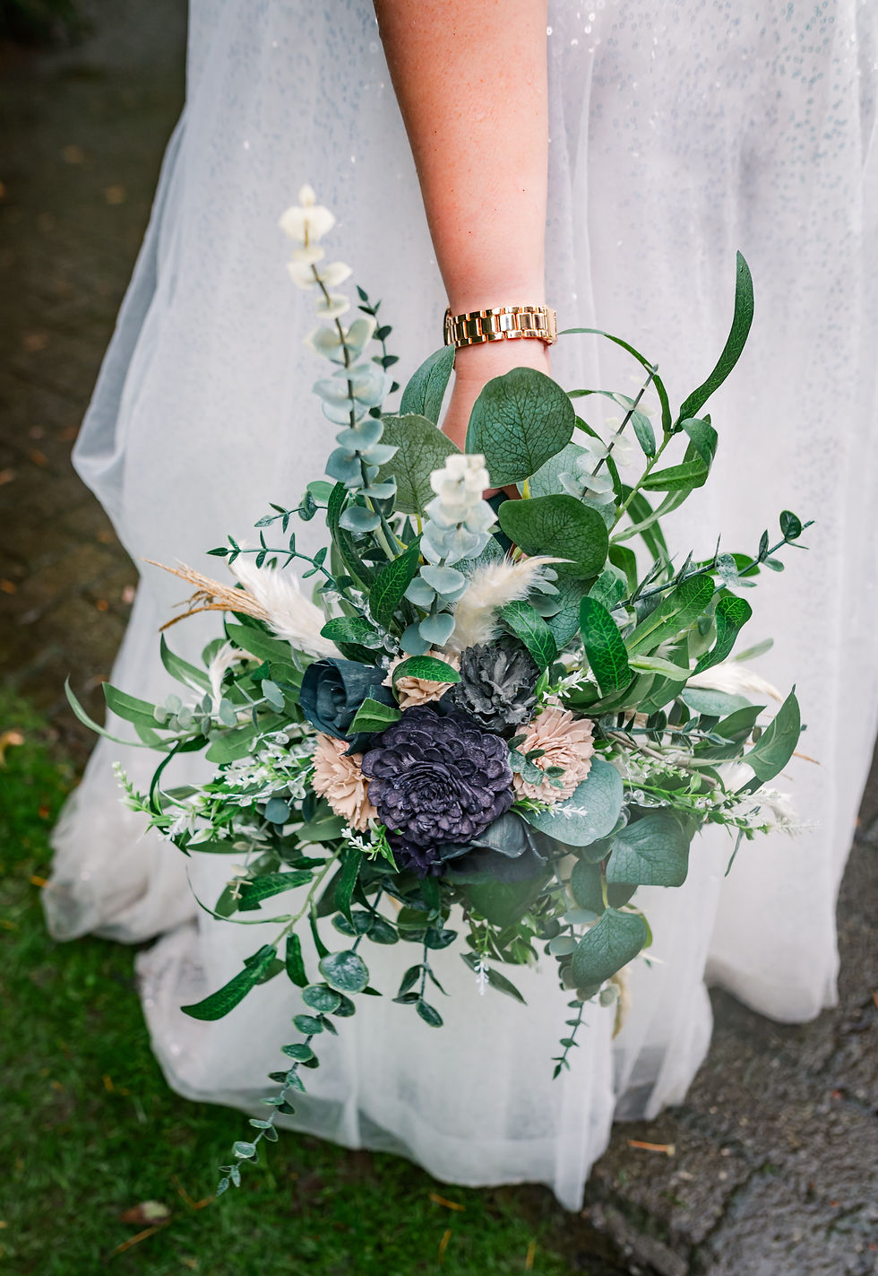 A person in a white dress holds a lush bouquet of green, white, and purple flowers, standing on a stone path with grass nearby.