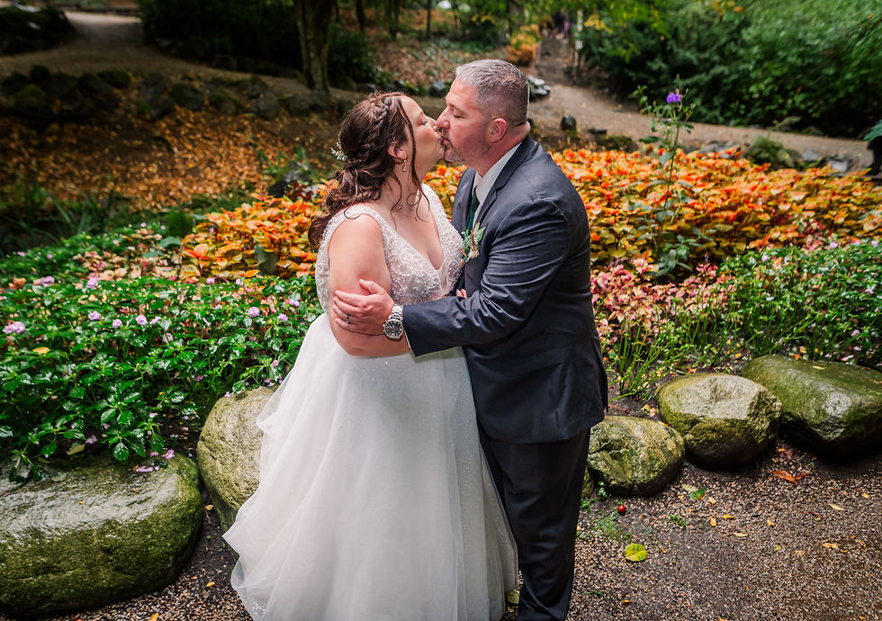 Bride and groom kissing in a lush garden, surrounded by colorful flowers and green foliage. Bride in white gown, groom in dark suit.