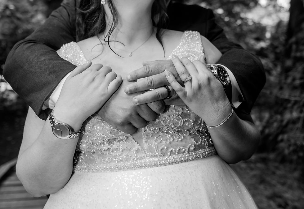 Bride and groom embrace, hands intertwined, showcasing rings and watches. Bride’s beaded gown visible. Romantic, woodland setting. Black and white.