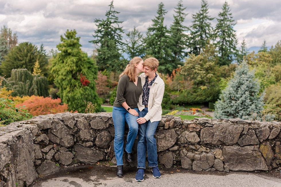 Couple sitting on a stone wall, smiling at each other in a vibrant park with colorful trees and cloudy sky in the background.