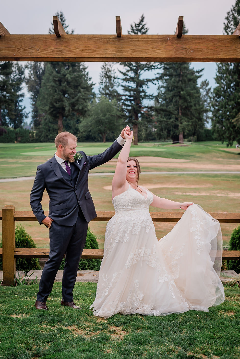 Bride and groom dancing joyfully outdoors on a golf course. Bride in lace gown, groom in suit. Wooden pergola and trees in the background.