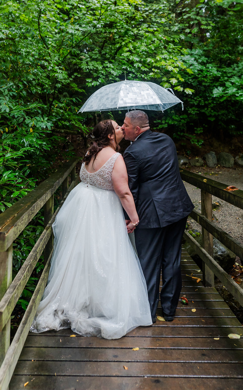 Bride and groom kiss under a clear umbrella on a wooden bridge, surrounded by lush green foliage. Bride in white dress, groom in black suit.