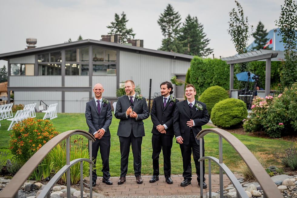 Four men in suits with boutonnieres stand smiling on a garden bridge. Background shows a modern building, greenery, and chairs. Festive mood.